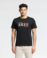 A man wears the Psycho Bunny MENS OCEANIA EMBROIDERED GRAPHIC TEE (B6U607F200) in black, showcasing a row of colorful abstract bunnies across the chest with retro vibes, paired with light pants.