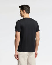A man with short brown hair wears a Psycho Bunny Men's Oceania Embroidered Graphic Tee - B6U607F200 in black and beige shorts, standing against a neutral backdrop, evoking subtle retro vibes.