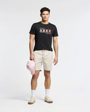 A man stands wearing a Psycho Bunny MENS OCEANIA EMBROIDERED GRAPHIC TEE - B6U607F200, giving off retro vibes. He pairs it with beige shorts, white socks, matching sneakers, and holds a pink cap in his right hand against a plain backdrop.
