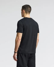 A man with short hair wears a Psycho Bunny MENS GOLD COAST GRAPHIC TEE - B6U631F200 and black shorts. He stands facing away from the camera against a light gray background, hands relaxed by his sides.