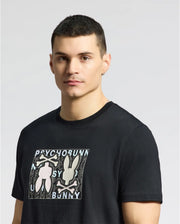 A person with short dark hair and an earring is wearing the Psycho Bunny MENS GOLD COAST GRAPHIC TEE - B6U631F200, a classic fit black Pima cotton T-shirt featuring two bunny silhouettes and "Psycho Bunny" text on a plain gray background.