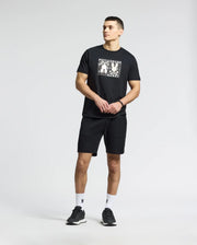 A man stands wearing Psycho Bunny's MENS GOLD COAST GRAPHIC TEE - B6U631F200 in black with a white graphic, paired with black shorts and sneakers. He gazes to the side with hands clasped against a plain, light gray background.