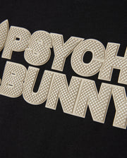 A close-up shot of the textured text "PSYCHO BUNNY" in large, bold, off-white letters on a black background. The wordmark graphic features a fine, grid-like pattern on the letters, providing a tactile, embossed look reminiscent of HD-silicone print. This detail is featured on the Psycho Bunny MENS MELVIN GRAPHIC TEE - B6U663C200.