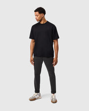 A person is standing against a plain grey background, wearing the MENS DUKE OVERSIZED TEE - B6U742D200 by Psycho Bunny, dark grey pants, and white sneakers. The relaxed fit t-shirt hangs comfortably as they look downward to their right with their arms by their sides.