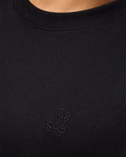 Close-up of a person wearing a Psycho Bunny MENS DUKE OVERSIZED TEE - B6U742D200, crafted from black heavyweight Pima cotton with a ribbed crew neckline. The relaxed fit tee features a subtle black embroidered chest bunny head with crossbones. The background is out of focus.