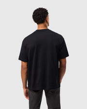 A person with short, curly hair is standing with their back to the camera. They are wearing a relaxed fit MENS DUKE OVERSIZED TEE - B6U742D200 by Psycho Bunny, made of heavyweight Pima cotton, paired with dark pants. The background is plain and light grey.