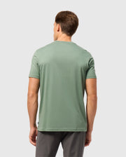 A man with short brown hair is standing with his back to the camera, wearing a green Psycho Bunny MENS MALTON GRAPHIC TEE - B6U795D200 and dark gray pants against a plain light gray background.