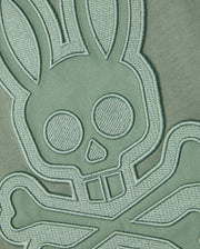A close-up of an embroidered patch on a Psycho Bunny MENS MALTON GRAPHIC TEE - B6U795D200 made from Pima cotton fabric. The design features a stylized skull with bunny ears and crossbones, detailed with stitching that raises it from the muted green background, giving it a three-dimensional look. Made in Peru, the unique appeal of this T-shirt is enhanced by its intricate craftsmanship.