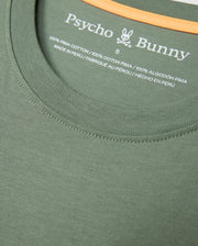Close-up of the collar and neckline area of the MENS MALTON GRAPHIC TEE - B6U795D200 in green by Psycho Bunny. The shirt has a small printed label displaying the brand name, logo, size, and material composition — 100% Pima Cotton, Made in Peru. An orange strip is visible inside the collar seam.