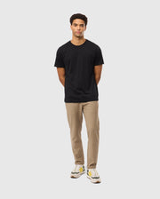 A man with short curly hair stands facing forward with his arms behind his back. He is wearing a Psycho Bunny MENS JAMES PREMIUM CREW NECK TEE - B6U952D200 made of Pima cotton, beige pants, and white sneakers with yellow and green accents, all against a plain gray background.
