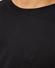 A close-up of a person showcasing the MENS JAMES PREMIUM CREW NECK TEE - B6U952D200 by Psycho Bunny. This black Pima cotton shirt features an embroidered black logo on the chest, depicting a bunny head with crossbones underneath. The person's neck and part of the shoulder are visible.