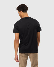 A man with short curly hair is standing with his back to the camera. He is wearing a black Psycho Bunny MENS JAMES PREMIUM CREW NECK TEE - B6U952D200 and light tan pants. The background is plain and light-colored.