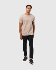 A person with short hair is standing against a plain background, wearing a beige Psycho Bunny MENS JAMES PREMIUM CREW NECK TEE - B6U952D200 made of premium Pima cotton, black pants, and black sneakers. They have a relaxed posture, one hand by their side and the other slightly bent, and they are looking directly at the camera.