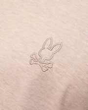 A close-up of the MENS JAMES PREMIUM CREW NECK TEE - B6U952D200 by Psycho Bunny in beige, crafted from premium Pima cotton, showcases a small, meticulously embroidered design featuring a stylized bunny head above two crossed bones, offering a playful twist on the classic skull and crossbones motif.
