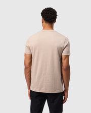 A person with short, curly hair is standing with their back to the camera, wearing a beige Psycho Bunny MENS JAMES PREMIUM CREW NECK TEE - B6U952D200 made from premium Pima cotton and black pants. The background is plain and light gray.