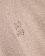 Close-up of the Psycho Bunny Men's James Premium Crew Neck Tee (B6U952D200) in premium Pima cotton, featuring an embroidered design of a bunny head on top of crossed bones, reminiscent of a skull and crossbones symbol. The meticulous embroidery is done in a slightly darker beige thread, offering a subtle contrast from the fabric.