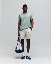 A man stands against a light background wearing the Psycho Bunny MENS JAMES PREMIUM CREW NECK TEE (B6U952D200) in light green, paired with beige shorts, white socks, blue sneakers, a blue cap, and holding a beige and navy tote bag.