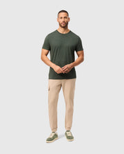 A man with short hair and a beard is wearing a stylish Psycho Bunny MENS JAMES PREMIUM CREW NECK TEE in dark green, made from soft cotton, beige cargo pants, and green sneakers. He is standing against a plain light grey background, looking at the camera with his hands clasped in front.