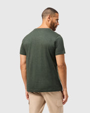 A man with short hair and a beard stands facing away from the camera. He is wearing a dark green, stylish *Psycho Bunny Men's James Premium Crew Neck Tee - B6U952D200* and light-colored pants. The background is plain and light-colored.