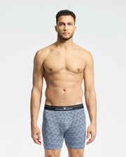 A man with short, curly hair stands against a plain backdrop, wearing Psycho Bunny's MENS 2 PACK BOXER BRIEF - B6V481F200. Crafted from a Pima cotton-Modal blend and accented by a black waistband, he gazes ahead neutrally, exuding all-day comfort.