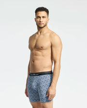 A man with short curly hair and facial hair stands against a plain background, wearing Psycho Bunny's MENS 2 PACK BOXER BRIEF - B6V481F200, made from a Pima cotton-Modal blend with a black waistband, exuding all-day comfort as his hands rest at his sides.