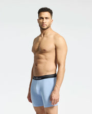 A man stands against a plain background, wearing Psycho Bunny's MENS 2 PACK BOXER BRIEF - B6V481F200, featuring light blue Pima cotton and a black waistband. He looks towards the camera with a neutral expression.