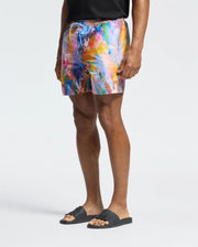 A person showcases vacation style in Psycho Bunny MENS MILOS PRINTED SWIM TRUNK with an abstract design, paired with a black shirt and slide sandals, set against a white background.