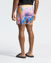 A person is standing, facing away, in Psycho Bunny's colorful MENS MILOS PRINTED SWIM TRUNK - B6W446F200 with an abstract pattern featuring small white symbols, paired with a black T-shirt and slides. The plain light gray background perfectly showcases their vacation style.