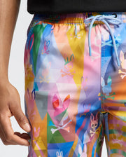 Wearing Psycho Bunny's MENS MILOS PRINTED SWIM TRUNK (B6W446F200), a person showcases colorful trunks with multicolored bunny and skull motifs and a light blue drawstring. Paired with a black shirt, these quick-dry shorts embody vacation style. Their hand appears to the left.