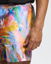 A person dons Psycho Bunny's MENS MILOS PRINTED SWIM TRUNK - B6W446F200, featuring a quick-dry, geometric design with multicolored skulls and bunny ears in vibrant pink, blue, green, and yellow. Perfect for vacation style, they stand with one hand by their side.