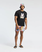A man displays his vacation style with a black t-shirt featuring a colorful bunny design, Psycho Bunny's MENS MILOS PRINTED SWIM TRUNK, and a multicolored bucket hat. Black slide sandals complete the look against a plain white backdrop.