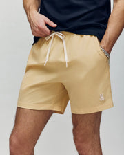 A person wears Psycho Bunny MENS SAMOS HYDROCHROMIC PRINTED SWIM TRUNK (B6W453F200) in light beige, featuring a small embroidered bunny logo on the left leg, paired with a dark t-shirt and their left hand in their pocket.