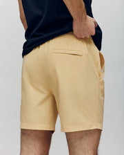 A person shown from the back wears Psycho Bunny’s MENS SAMOS HYDROCHROMIC PRINTED SWIM TRUNK - B6W453F200 in light beige, featuring a subtle bunny pattern, set against a plain background. One hand is in a pocket; the other touches the waistband.
