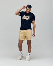 A man wearing a black graphic t-shirt, Psycho Bunny MENS SAMOS HYDROCHROMIC PRINTED SWIM TRUNK, white socks, and white sneakers adjusts his sunglasses with one hand while looking down against a plain background.