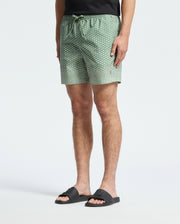A person wears Psycho Bunny's MENS BENDIGO ALL OVER PRINTED SWIM TRUNK and black sliders. The green polka dot shorts have a drawstring, and the person stands against a plain white background with a partially visible black shirt.