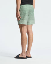 A person is wearing Psycho Bunny's MENS BENDIGO ALL OVER PRINTED SWIM TRUNK (B6W619F200) and a black t-shirt. The quick-dry fabric whispers adventure as they stand with their back to the camera, complemented by black slides against a plain, light-colored background.