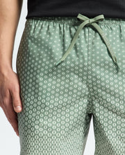 A person wears Psycho Bunny's MENS BENDIGO ALL OVER PRINTED SWIM TRUNK - B6W619F200, featuring a green leaf pattern and drawstring waistband. Their hand rests by their side, paired with a black top against a light background.