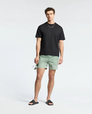 A person in a Psycho Bunny black t-shirt and MENS BENDIGO ALL OVER PRINTED SWIM TRUNK (B6W619F200) with a quirky bunny pattern, black sandals, sunglasses in hand, curly hair, and a gold chain necklace poses against a plain white background.