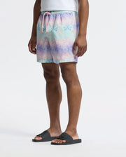 A person is wearing Psycho Bunny's pastel bunny-printed swim trunk (MENS FORSTER BUNNY PRINTED SWIM TRUNK - B6W621F200) against a plain background. They complement it with black slide sandals for a relaxed, casual style.
