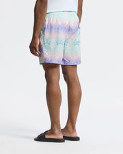 A person with medium brown skin wearing pastel-colored tie-dye Psycho Bunny Men's Forster Bunny Printed Swim Trunks (B6W621F200) and black slide sandals stands against a plain background, facing away. The shorts' purple, blue, and green hues evoke an ocean view.