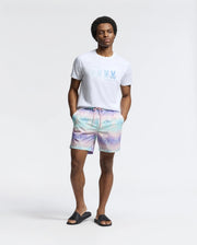 Wearing a Psycho Bunny MENS FORSTER BUNNY PRINTED SWIM TRUNK - B6W621F200, the man exudes effortless cool in pastel tie-dye shorts and black slide sandals, posing with hands in pockets against a white background.