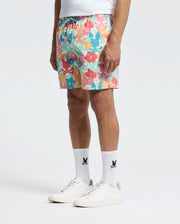 A person exemplifies comfort and style wearing Psycho Bunny's Lemmon All Over Print Swim Trunk, featuring vibrant pink, blue, and yellow floral patterns. They pair it with logo-adorned white socks and sneakers against a plain white backdrop.