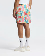 A person wearing Psycho Bunny's MENS LEMMON ALL OVER PRINT SWIM TRUNK with a white shirt and socks featuring black logos pairs them with white sneakers. The polyester mechanical stretch fabric offers comfort and style, all set against a plain white background.