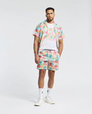 A man in a Psycho Bunny MENS LEMMON ALL OVER PRINT SWIM TRUNK, colorful floral shirt, matching shorts, and white T-shirt stands against a plain background. Completed with socks and sneakers, the polyester mechanical stretch outfit embodies comfort and style for ease of movement.