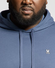 A close-up shows a person wearing the Psycho Bunny MENS BIG AND TALL LEVY ESSENTIAL FLEECE HOODIE, featuring a small white bunny and crossbones logo on the chest. The hoodie is blue, and part of the person's face and beard are visible.