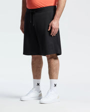 A person stands against a plain white background wearing Psycho Bunny's MENS BIG AND TALL LEVANZO EMBROIDERED MICRO FRENCH TERRY SWEATSHORT (B9R578F200) in black with a small bunny logo, matching white bunny-logo socks, white sneakers, and an orange shirt.