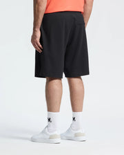 A person stands against a plain white background, seen from the back, wearing a coral shirt, white sneakers, white socks with black Adidas logos, and Psycho Bunny MENS BIG AND TALL LEVANZO EMBROIDERED MICRO FRENCH TERRY SWEATSHORTS.