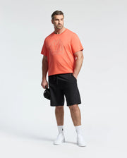 A man stands against a white background wearing a bright coral graphic t-shirt, Psycho Bunny MENS BIG AND TALL LEVANZO EMBROIDERED MICRO FRENCH TERRY SWEATSHORT in black, white socks and sneakers, holding a black cap in his left hand.
