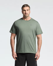 A man wears a sage green BIG AND TALL CLASSIC CREW NECK TEE from Psycho Bunny, made of 100% Pima cotton and featuring a small embroidered chest logo. He pairs it with black shorts, standing against a white background, sporting short hair and a beard.