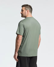A bearded man with short hair stands back turned, displaying the green Psycho Bunny Big and Tall Classic Crew Neck Tee (B9U014CRPC). Paired with black shorts, his stylish look contrasts against a plain white backdrop.